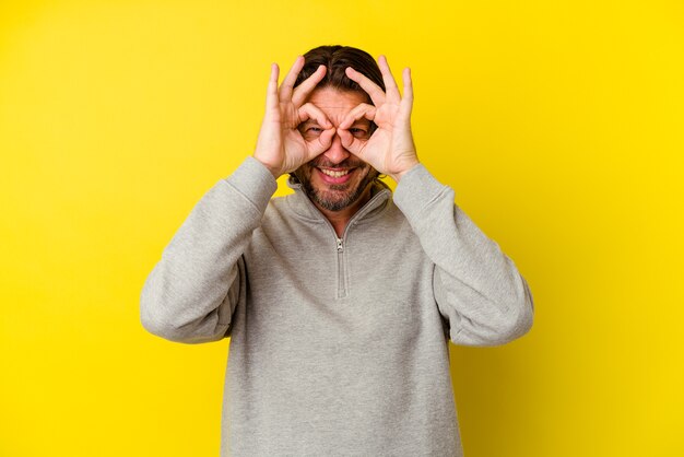 Middle age caucasian man isolated on yellow background excited keeping ok gesture on eye.