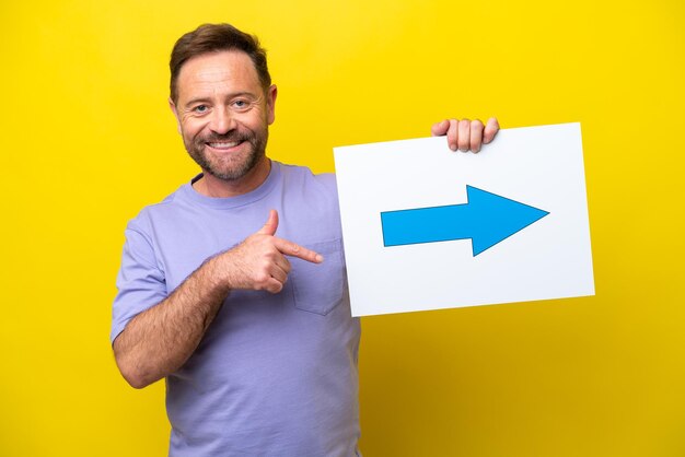 Photo middle age caucasian man isolated on yellow background holding a placard with arrow symbol and pointing it