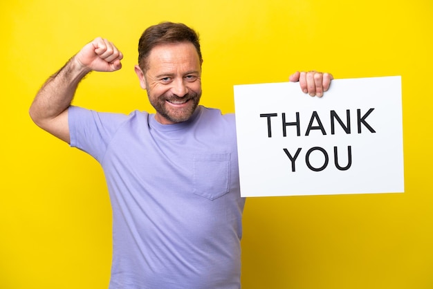 Photo middle age caucasian man isolated on yellow background holding a placard with text thank you and doing strong gesture