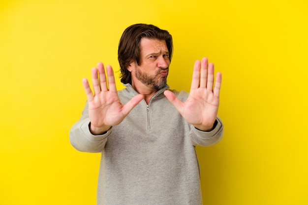 Middle age caucasian man isolated on yellow wall standing with outstretched hand showing stop sign, preventing you.