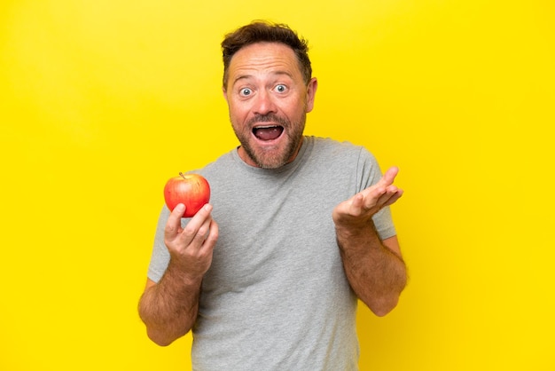 Middle age caucasian man with an apple isolated on yellow background with shocked facial expression