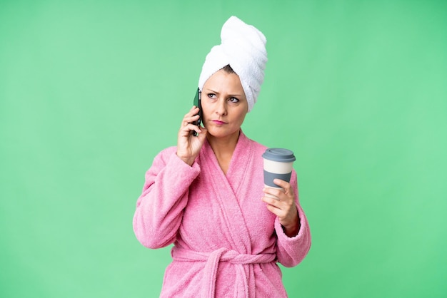 middle age caucasian woman in a bathrobe over isolated background holding coffee to take away and a mobile