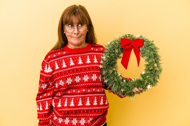 黄色の背景に分離された花輪を保持しているクリスマスを祝う中年の白人女性は混乱し、疑わしく、不安を感じています。