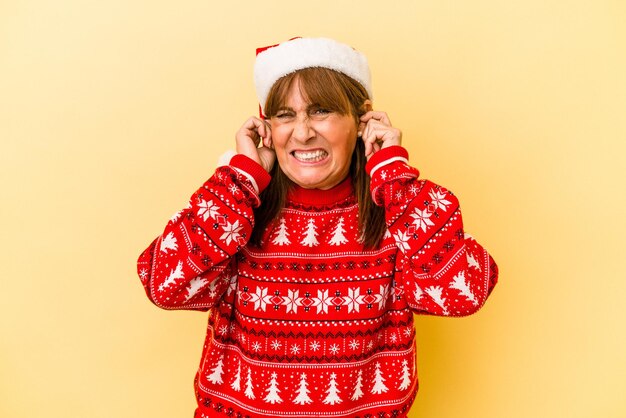 手で耳を覆う黄色の背景で隔離のクリスマスを祝う中年白人女性。