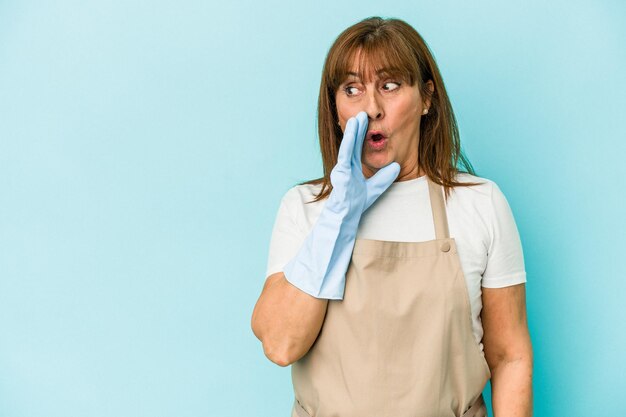 middle age caucasian woman cleaning home isolated on blue background is saying a secret hot braking news and looking aside