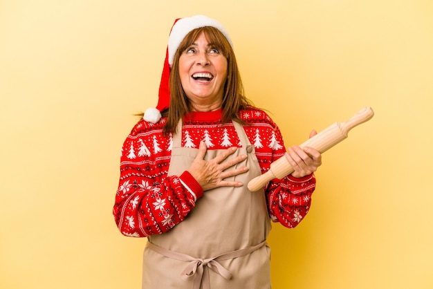 Middle age caucasian woman cooking cookies for christmas isolated on yellow background laughs out loudly keeping hand on chest.