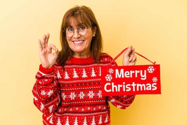 黄色の背景に分離された「メリークリスマス」のプラカードを持っている中年の白人女性は、陽気で自信を持って大丈夫なジェスチャーを示しています。