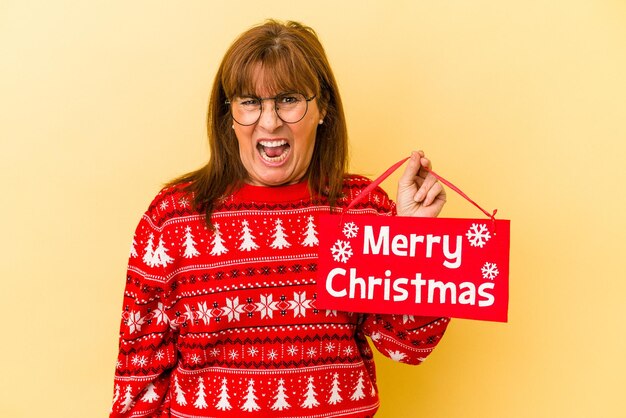 黄色の背景に分離された「メリークリスマス」のプラカードを持っている中年の白人女性は、非常に怒って攻撃的な叫び声を上げます。