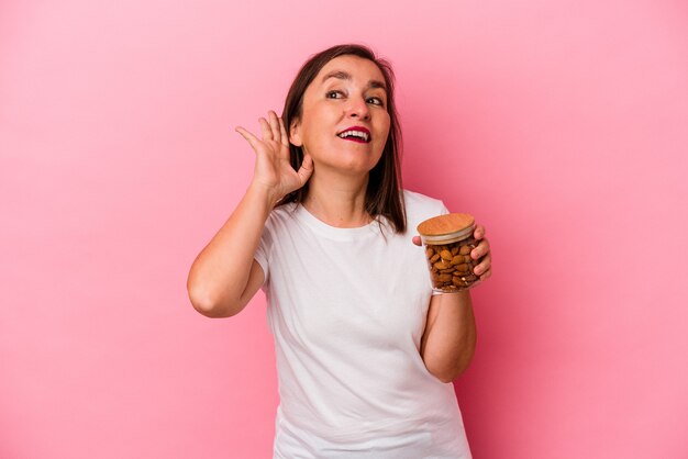 Middle age caucasian woman holding an almond jar isolated on pink background trying to listening a gossip.