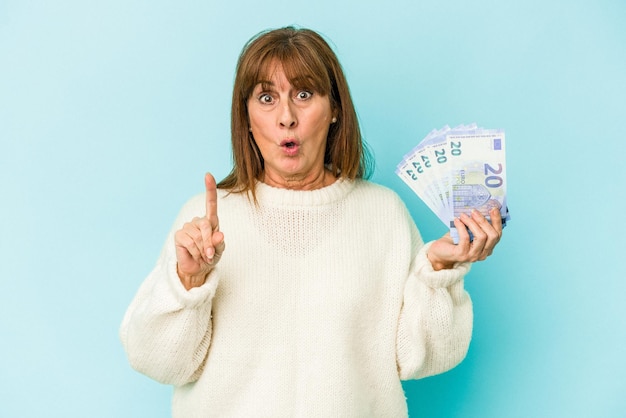 Middle age caucasian woman holding bank notes isolated on blue background having some great idea, concept of creativity.