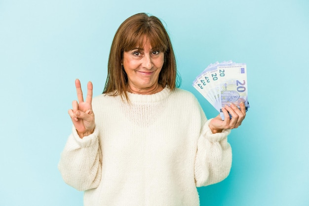 Middle age caucasian woman holding bank notes isolated on blue background showing number two with fingers.