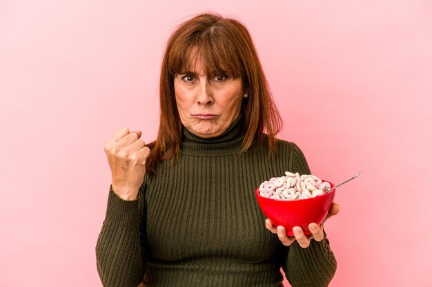 Middle age caucasian woman holding bowl of cereals isolated on pink background showing fist to camera, aggressive facial expression.