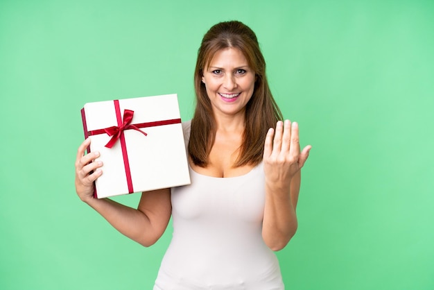 Middle age caucasian woman holding a gift over isolated background inviting to come with hand Happy that you came