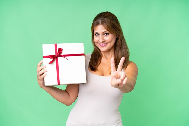 Middle age caucasian woman holding a gift over isolated background smiling and showing victory sign