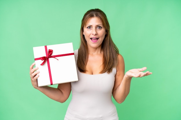 Middle age caucasian woman holding a gift over isolated background with shocked facial expression