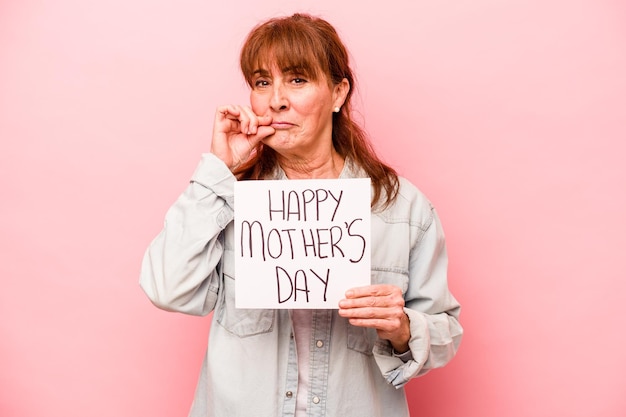 Кавказская женщина средних лет держит плакат с Днем матери на розовом фоне с пальцами на губах, хранящими секрет