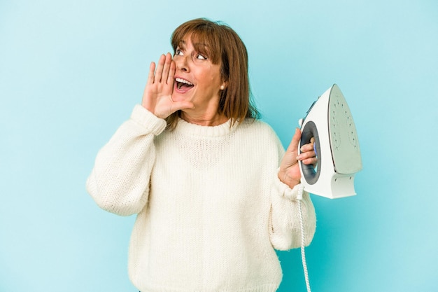 Middle age caucasian woman holding a iron isolated on blue background shouting and holding palm near opened mouth.