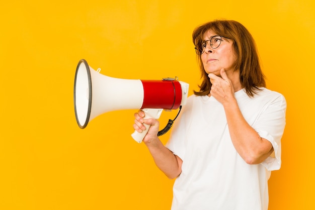 Middle age caucasian woman holding a megaphone looking sideways with doubtful and skeptical expression.