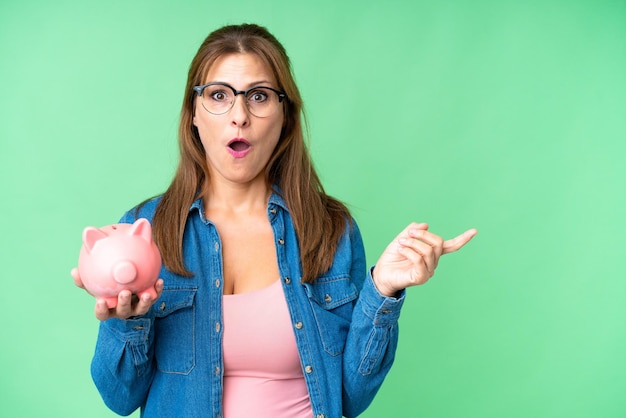 Middle age caucasian woman holding a piggybank over isolated background surprised and pointing side