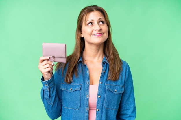 Middle age caucasian woman holding wallet over isolated background looking up while smiling
