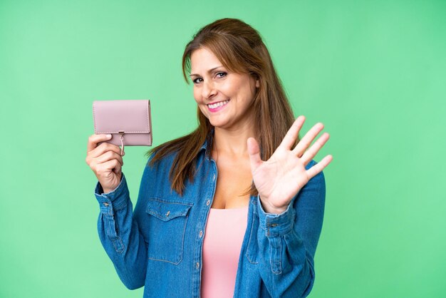 Middle age caucasian woman holding wallet over isolated background saluting with hand with happy expression