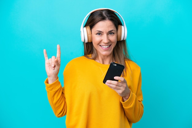 Middle age caucasian woman isolated on blue background listening music with a mobile making rock gesture