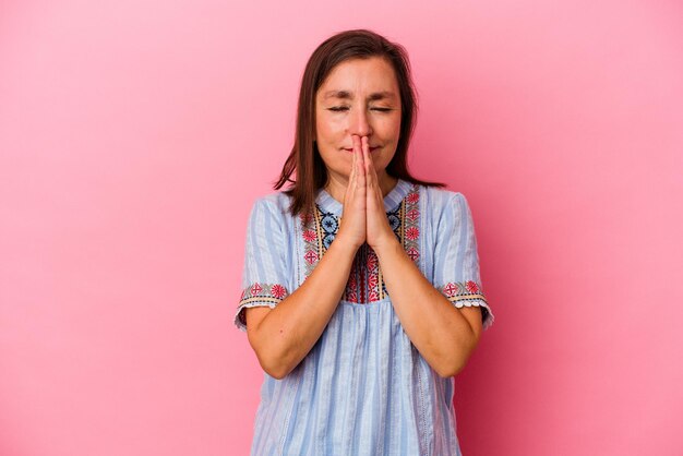 Middle age caucasian woman isolated on pink background holding hands in pray near mouth, feels confident.