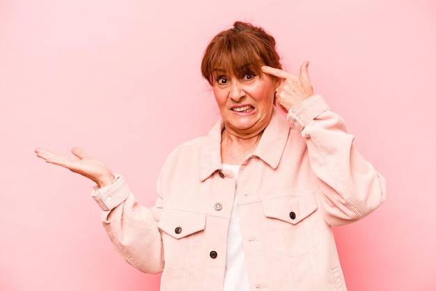 Middle age caucasian woman isolated on pink background holding and showing a product on hand