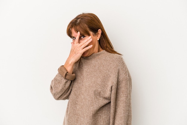 Middle age caucasian woman isolated on white background blink at the camera through fingers, embarrassed covering face.