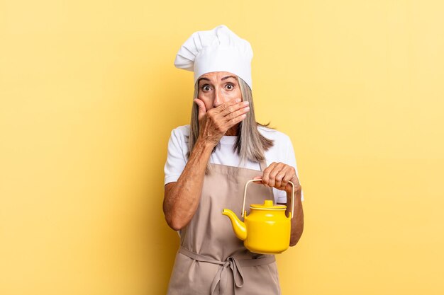 Middle age chef woman covering mouth with hands with a shocked, surprised expression, keeping a secret or saying oops and holding a teapot
