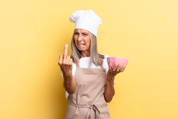 Middle age chef woman feeling angry, annoyed, rebellious and aggressive, flipping the middle finger, fighting back and holding an empty pot