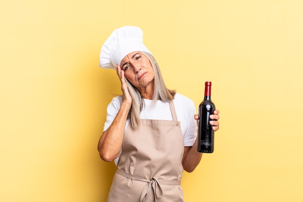 Donna chef di mezza età che si sente annoiata, frustrata e assonnata dopo un compito noioso, noioso e noioso, tenendo il viso con la mano che tiene una bottiglia di vino