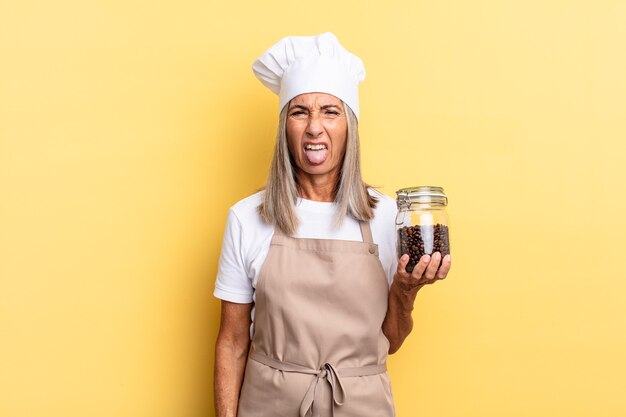 Middle age chef woman feeling disgusted and irritated, sticking tongue out, disliking something nasty and yucky holding coffee beans