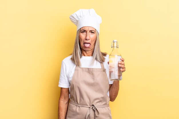 Middle age chef woman feeling disgusted and irritated, sticking tongue out, disliking something nasty and yucky with a water bottle