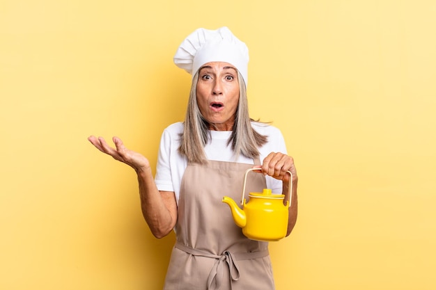 Middle age chef woman feeling extremely shocked and surprised, anxious and panicking, with a stressed and horrified look and holding a teapot