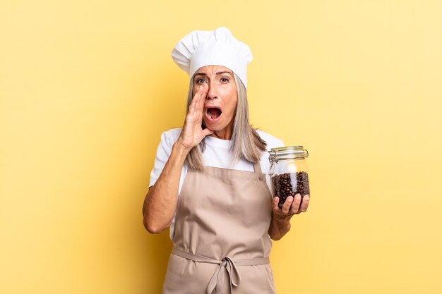 Middle age chef woman feeling happy, excited and positive, giving a big shout out with hands next to mouth, calling out holding coffee beans