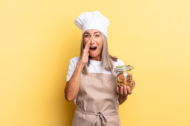 Middle age chef woman feeling happy excited and positive giving a big shout out with hands next to mouth calling out with cookies