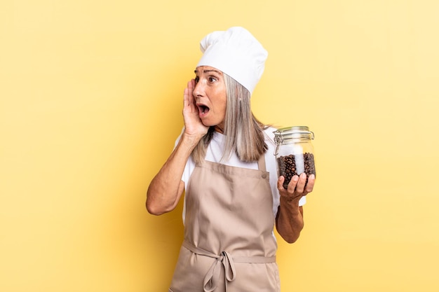 Middle age chef woman feeling happy, excited and surprised, looking to the side with both hands on face holding coffee beans