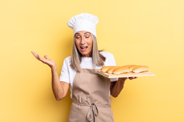 Middle age chef woman feeling happy, excited, surprised or shocked, smiling and astonished at something unbelievable and holding a bread tray