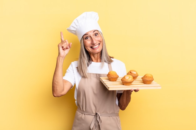 Middle age chef woman feeling like a happy and excited genius after realizing an idea, cheerfully raising finger, eureka! and holding a muffins tray