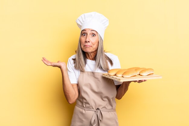 Donna chef di mezza età che si sente perplessa e confusa, dubitando, ponderando o scegliendo diverse opzioni con un'espressione divertente e tenendo in mano un vassoio del pane