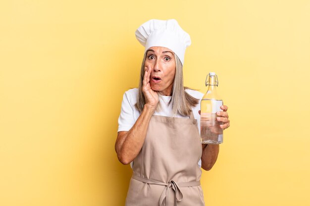 Middle age chef woman feeling shocked and scared, looking terrified with open mouth and hands on cheeks with a water bottle