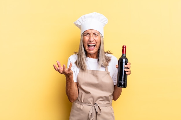 Middle age chef woman looking angry, annoyed and frustrated screaming wtf or whatÃ¢ÂÂs wrong with you holding a wine bottle