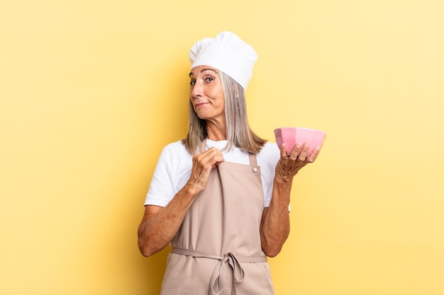 Foto donna chef di mezza età che sembra arrogante, di successo, positiva e orgogliosa, indicando se stessa e tenendo una pentola vuota