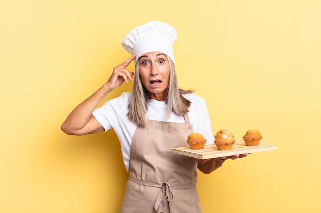 Donna chef di mezza età che sembra felice, stupita e sorpresa, sorride e realizza incredibili e incredibili buone notizie e tiene in mano un vassoio di muffin