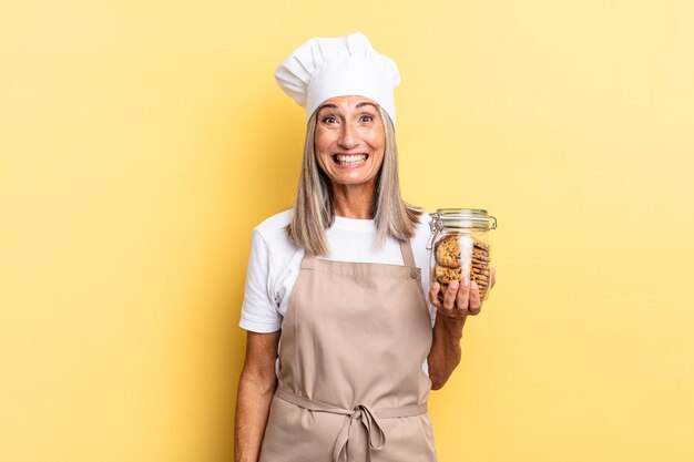 Middle age chef woman looking happy and pleasantly surprised excited with a fascinated and shocked expression with cookies