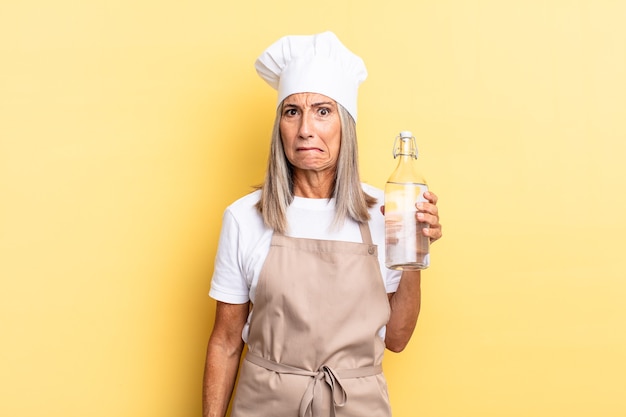 Middle age chef woman looking puzzled and confused, biting lip with a nervous gesture, not knowing the answer to the problem with a water bottle