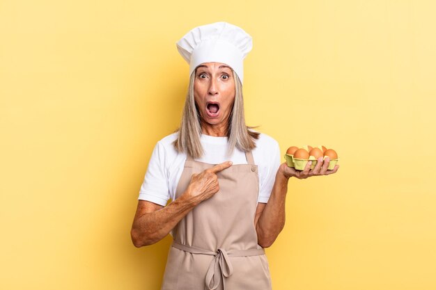 Middle age chef woman looking shocked and surprised with mouth wide open, pointing to self holding an eggs box