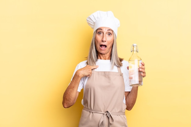 Middle age chef woman looking shocked and surprised with mouth wide open, pointing to self with a water bottle