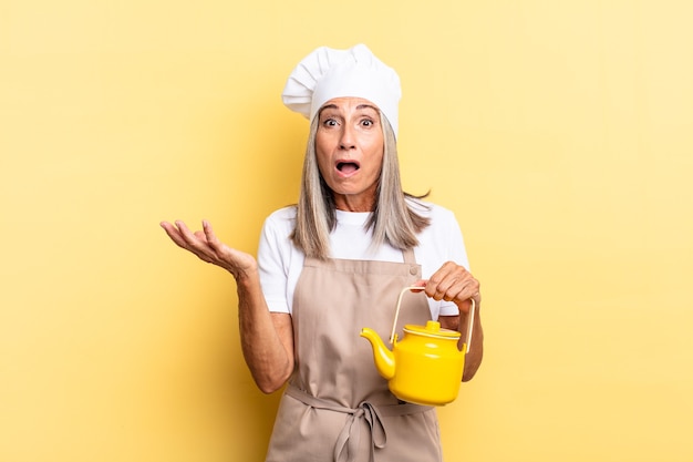 Middle age chef woman open-mouthed and amazed, shocked and astonished with an unbelievable surprise and holding a teapot